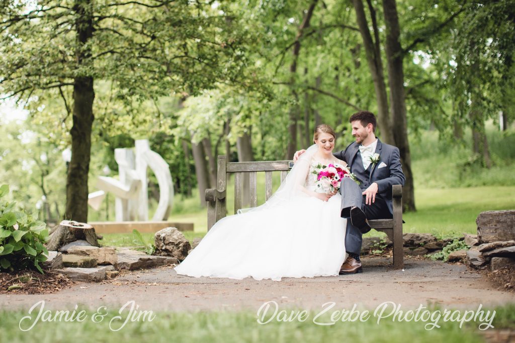 jamie and jim wedding photos sitting on a bench in a park with modern art. Photo captured by Dave Zerbe Photography.