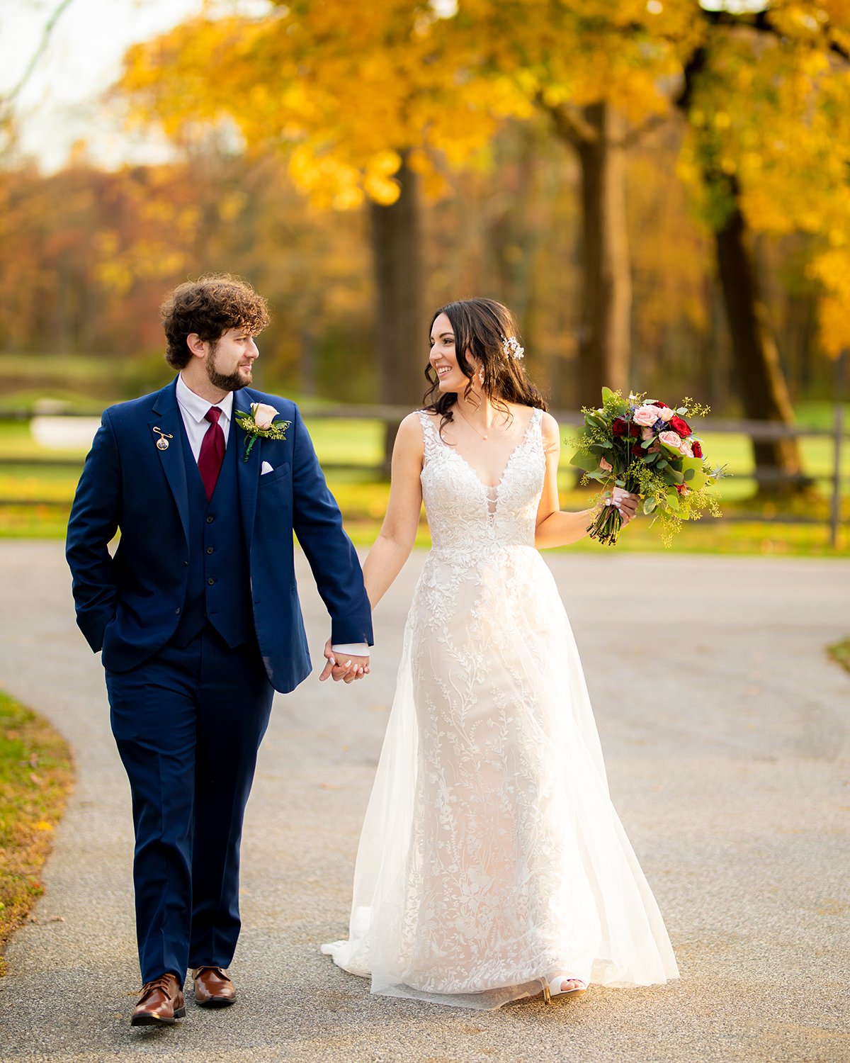 bride and groom holding hands as they walk down a road with yellow autumn trees behind them